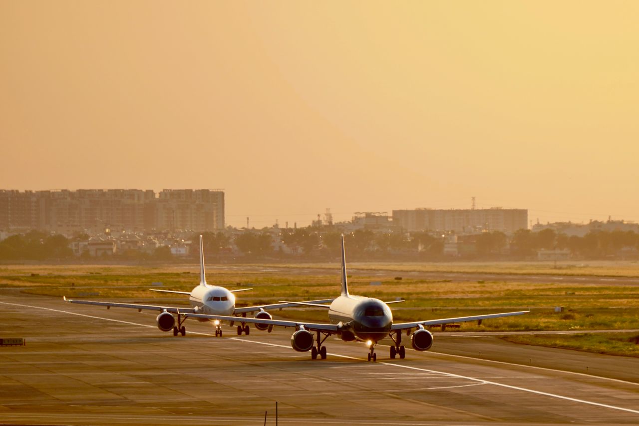 Two commercial airplanes taxiing at sunset with cityscape in background.